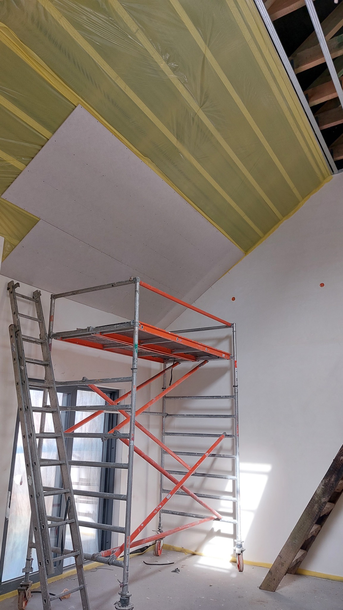Montaż płyty gipsowo-kartonowej na poddaszu z widocznym rusztowaniem, drabina i folia ochronna na izolacji. Elementy konstrukcyjne dachu widoczne w tle.