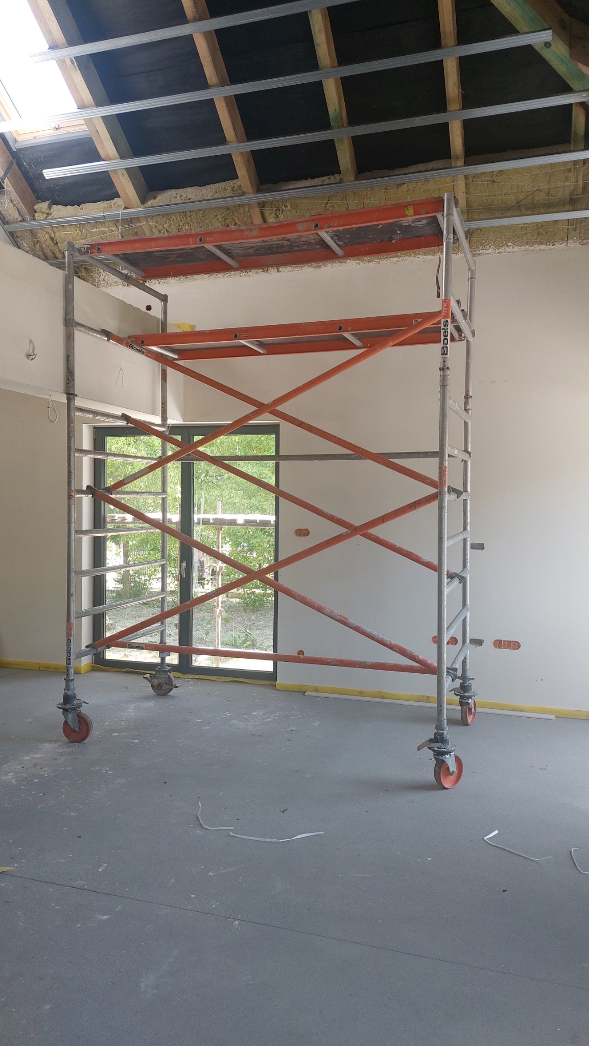 Wnętrze w trakcie prac wykończeniowych: rusztowanie na tle okna, ocieplone poddasze z widoczną konstrukcją metalową i drewnianą, surowa ściana z taśmą malarską przy podłodze.