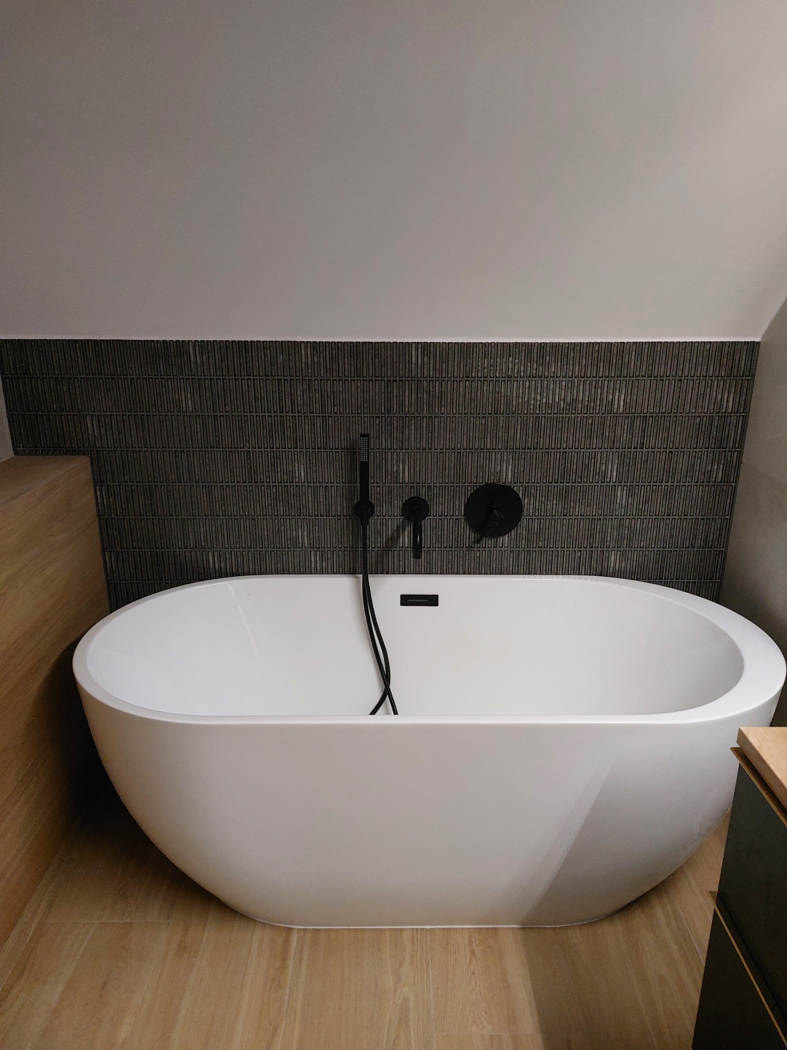 Nowoczesna łazienka z wolnostojącą białą wanną, czarna armatura i ściana wyłożona ciemnymi, pionowymi płytkami imitującymi drewno, oświetlenie górne.