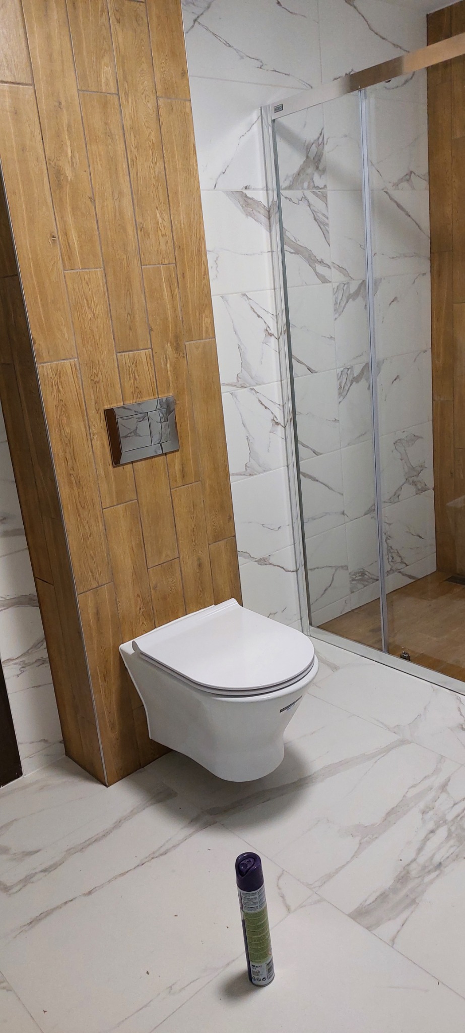 Nowoczesna toaleta podwieszana na drewnopodobnej ściance z ukrytym systemem spłukiwania, kabina prysznicowa z marmurowymi płytkami w tle, na podłodze stoi odświeżacz powietrza.