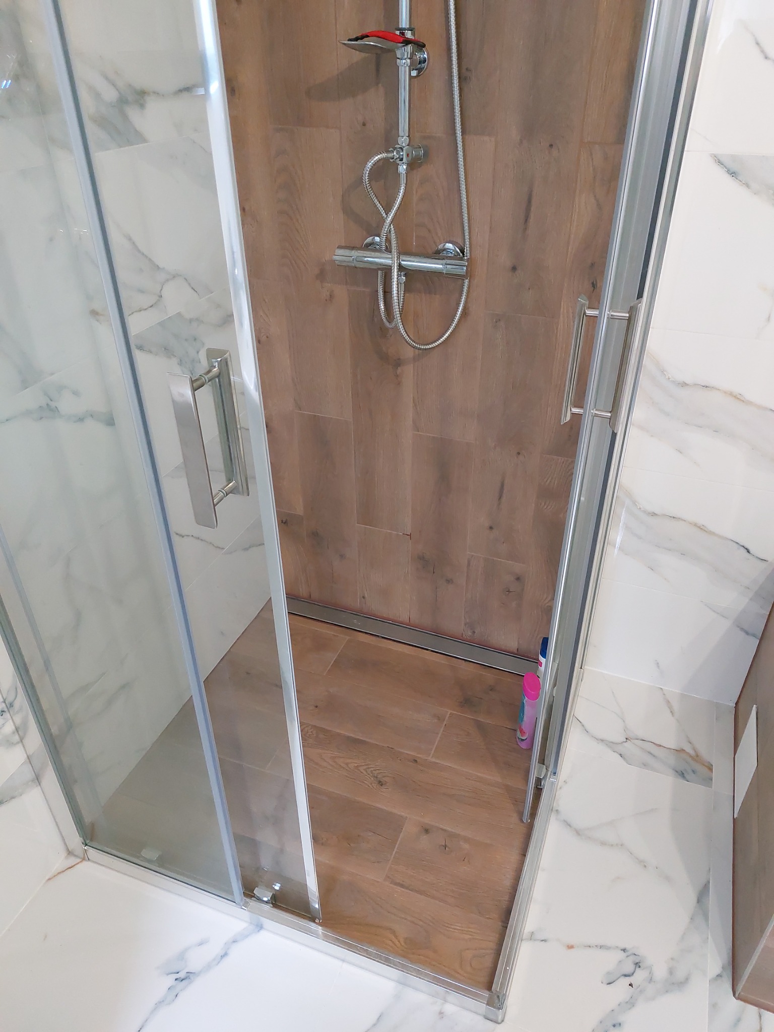 Kabina prysznicowa z przesuwnymi drzwiami, wykończona drewnopodobnymi płytkami wewnątrz i marmurowymi na zewnątrz, widoczny zestaw natryskowy i butelka kosmetyku w rogu.