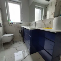 Nowoczesna łazienka z szafką pod umywalką w kolorze granatowym, marmurowymi płytkami na ścianach i podłodze oraz toaletą wiszącą.