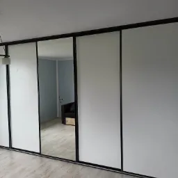 Nowoczesna szafa przesuwna z białymi frontami i czarnymi ramami, z lustrem odbijającym fragment pokoju z szarą sofą.