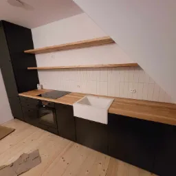 Nowoczesna kuchnia w odcieniach czerni i drewna, z białym ceramicznym zlewem farmerskim, drewnianym blatem i płytą indukcyjną, umieszczona na poddaszu ze skośnym sufitem i drewnianymi półkami.