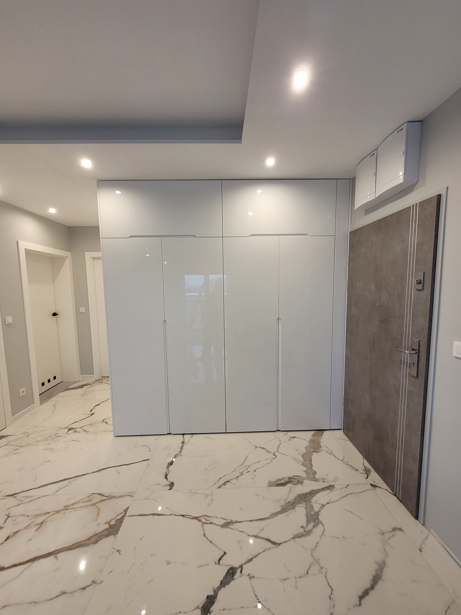Biała, wysoka szafa wnękowa z połyskiem, zintegrowana z sufitem, na podłodze z marmurowym wzorem, widoczna w przedpokoju mieszkania.