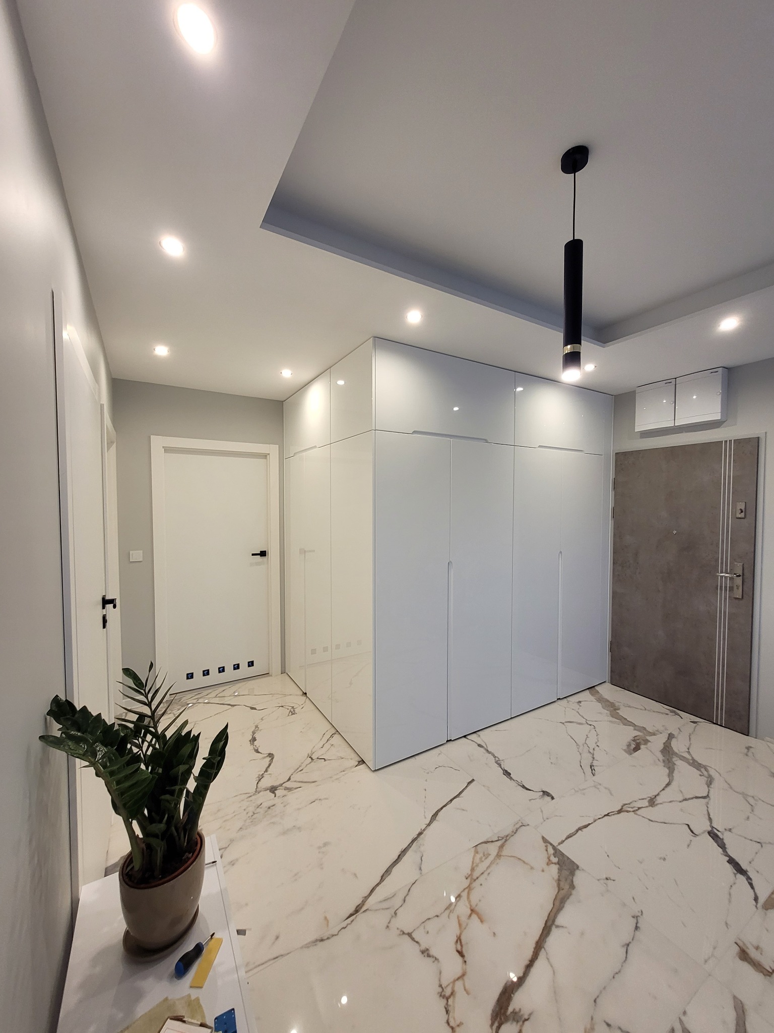 Biała, błyszcząca szafa wnękowa od podłogi do sufitu, zintegrowana z dwoma sąsiednimi drzwiami, na marmurowej podłodze z ciemnymi żyłami; w rogu widoczna roślina doniczkowa i narzędzia.