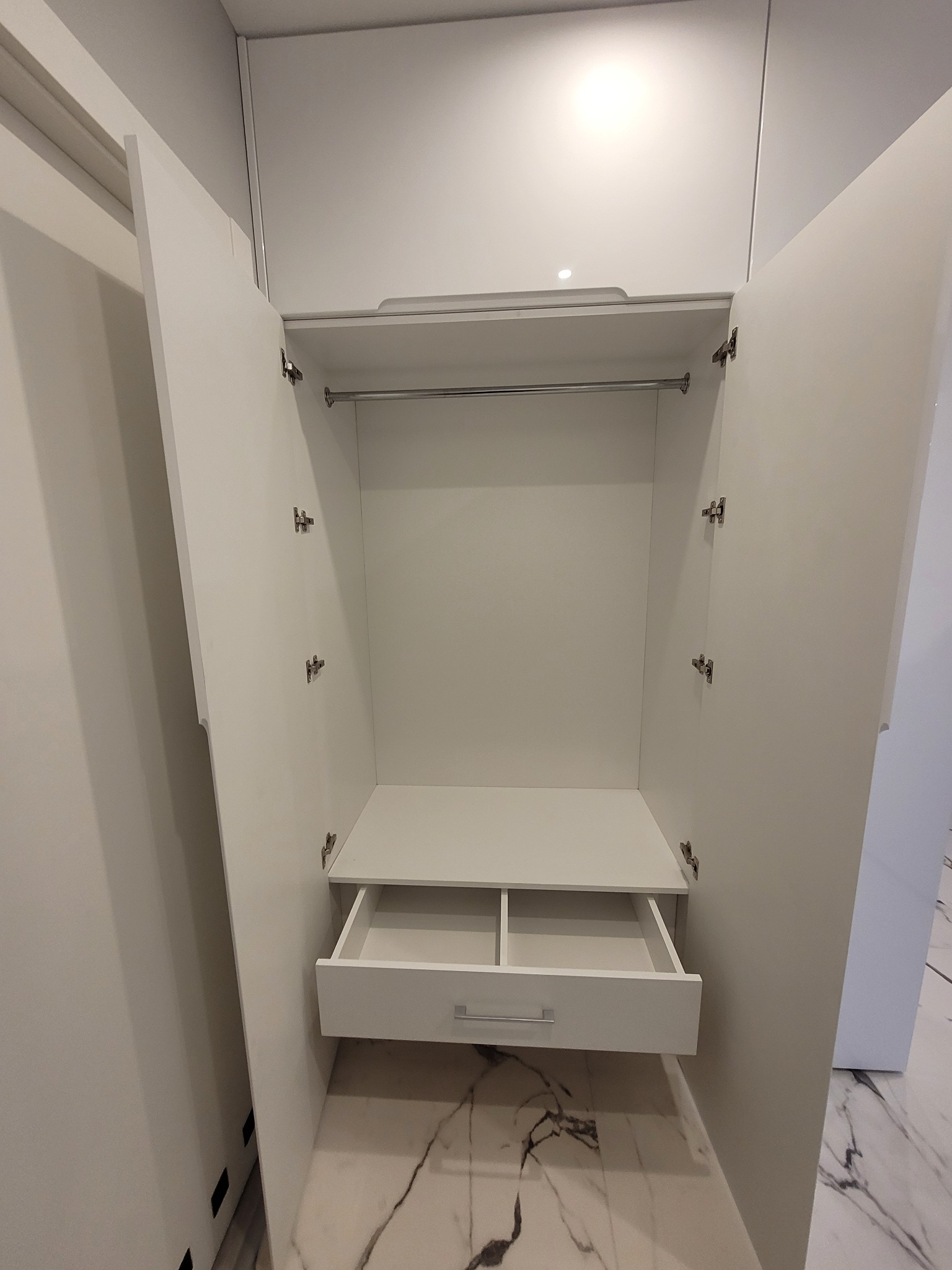 Wnętrze otwartej, białej szafy z wysuwaną szufladą i drążkiem na ubrania, widok na marmurową podłogę.