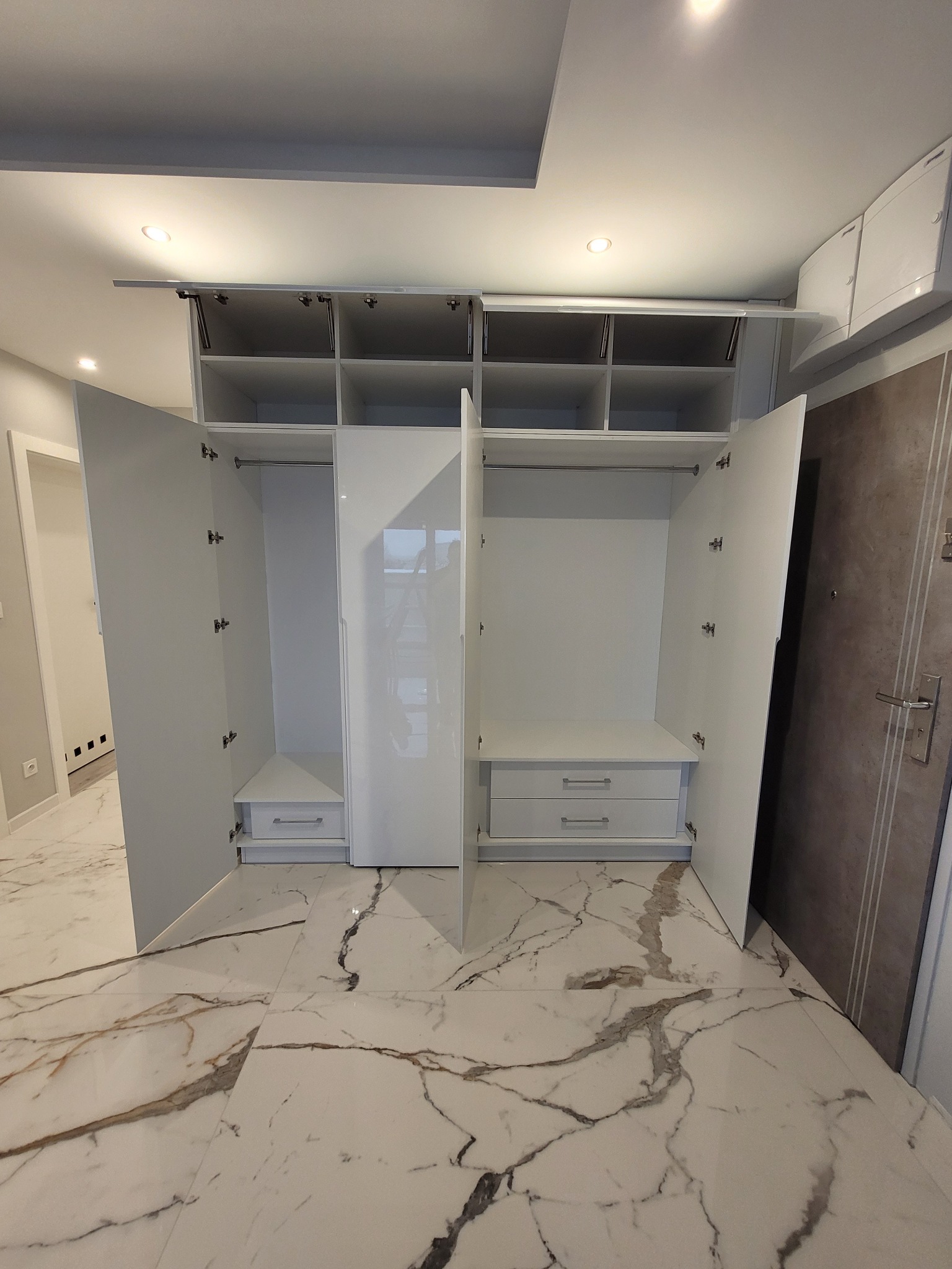 Biała, lakierowana szafa wnękowa z otwartymi drzwiami, widoczne półki, szuflady i drążek na wieszaki, na podłodze marmurowe płytki.