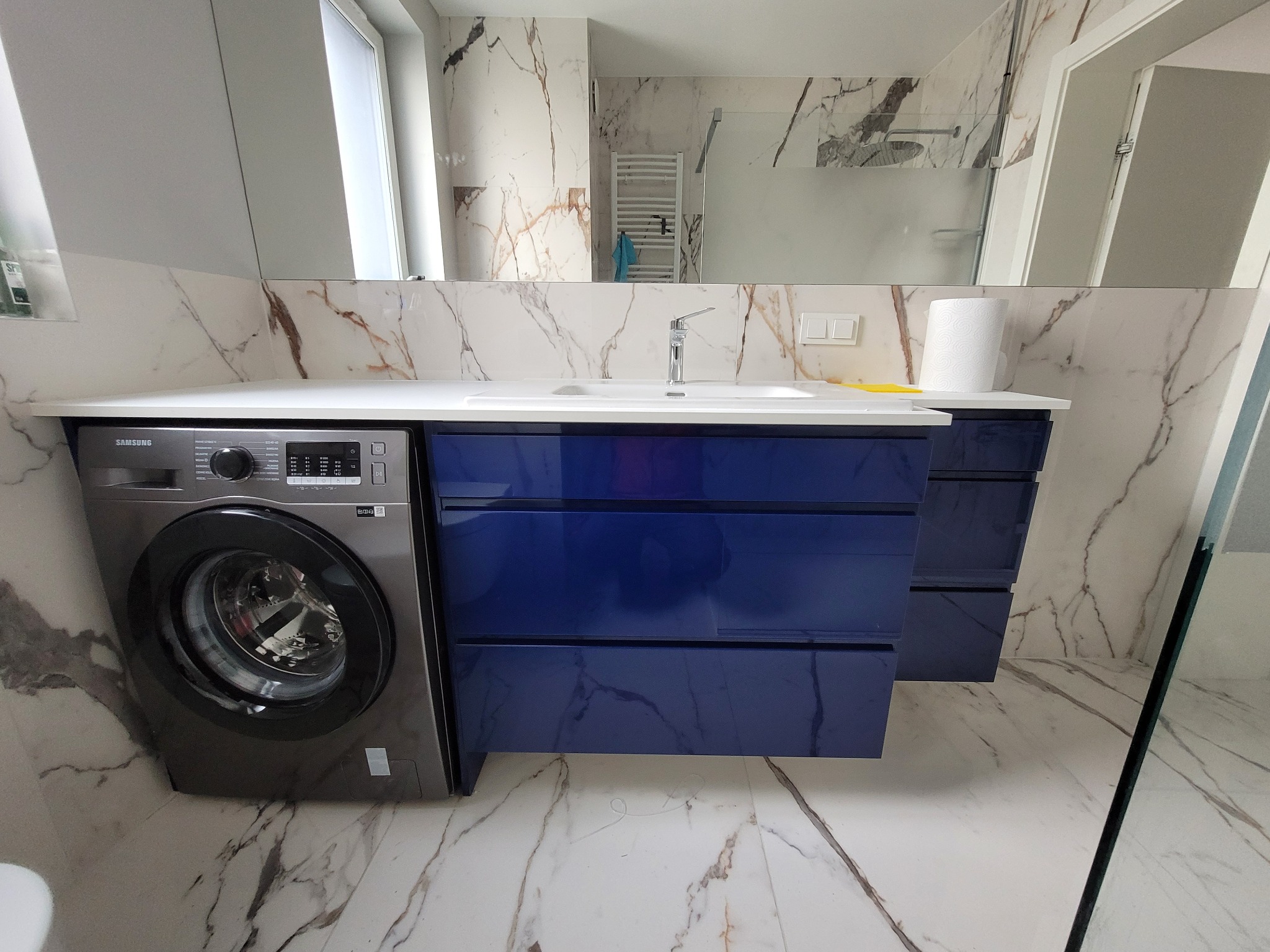 Nowoczesna łazienka z szafką na wymiar w kolorze granatowym, zintegrowaną z pralką Samsung i blatem z umywalką, na tle marmurowych płytek.