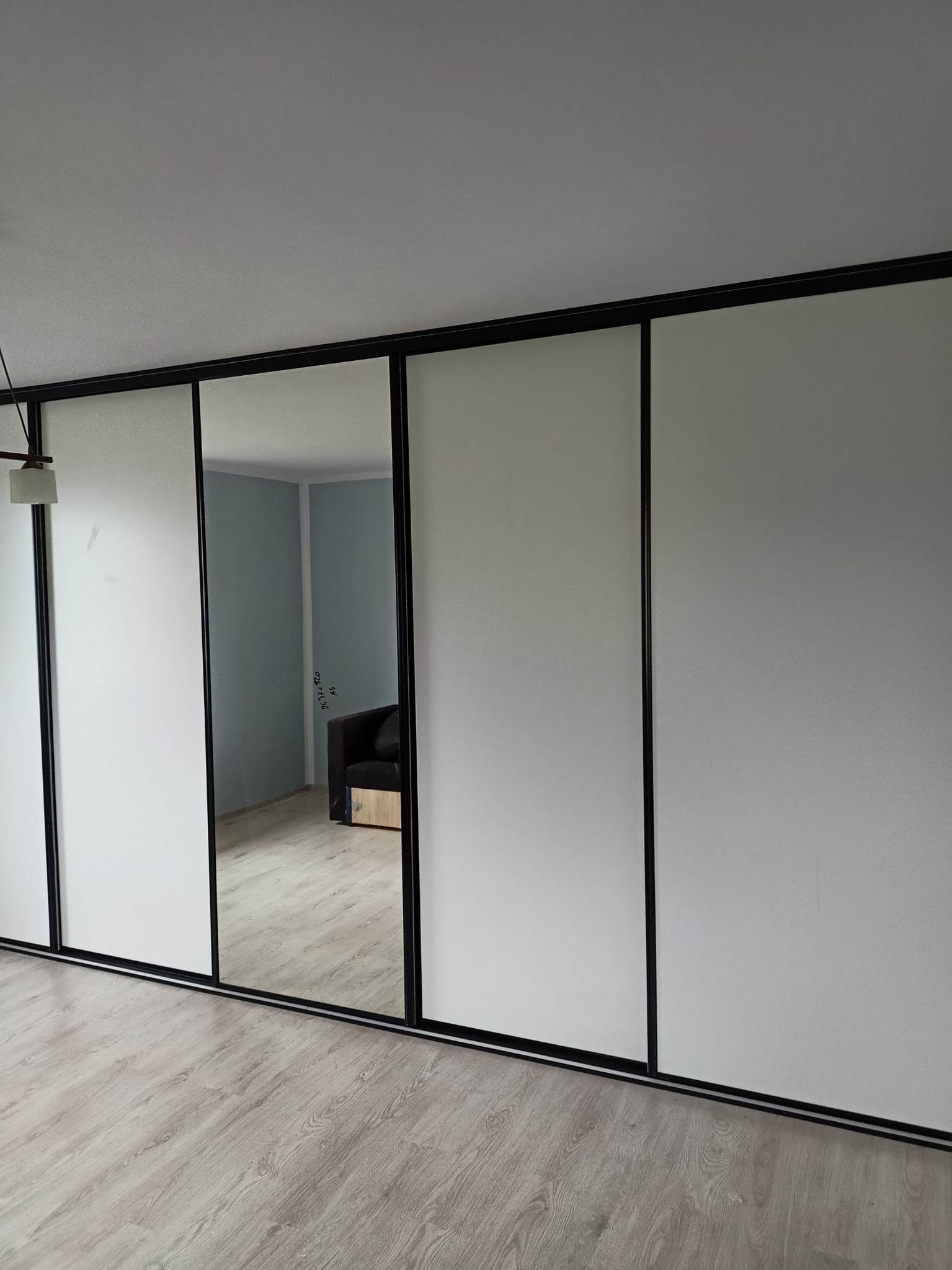 Nowoczesna szafa przesuwna z białymi frontami i czarnymi ramami, z lustrem odbijającym fragment pokoju z szarą sofą.