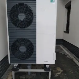 Biała pompa ciepła Vaillant aroTHERM, zamontowana na szarej, metalowej konstrukcji, z widocznym odpływem, umieszczona na brukowanej powierzchni przy ścianie budynku.