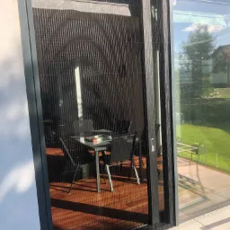 Zasłonięte drzwiami przesuwnymi z siatką plisowaną, widok na stół i krzesła na drewnianym tarasie oraz trawnik w tle.