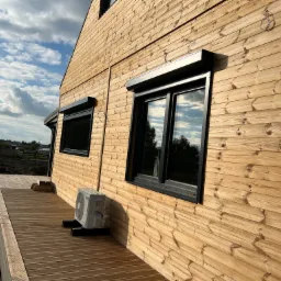 Elewacja domu obłożona drewnem z zamontowanymi oknami z czarnymi ramami i zwiniętymi roletami w kolorze czarnym, widoczny klimatyzator na drewnianym tarasie