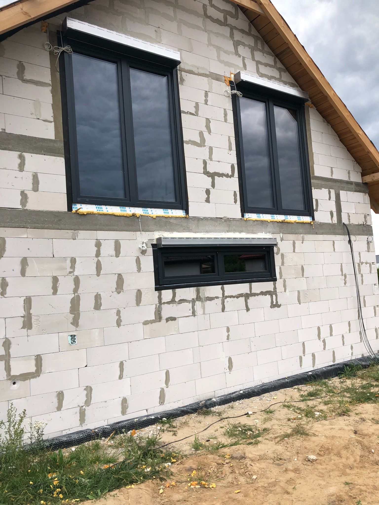 Surowy stan budynku z zamontowanymi oknami i roletami zewnętrznymi, widoczne przewody i pianka montażowa, ściana z białego gazobetonu.