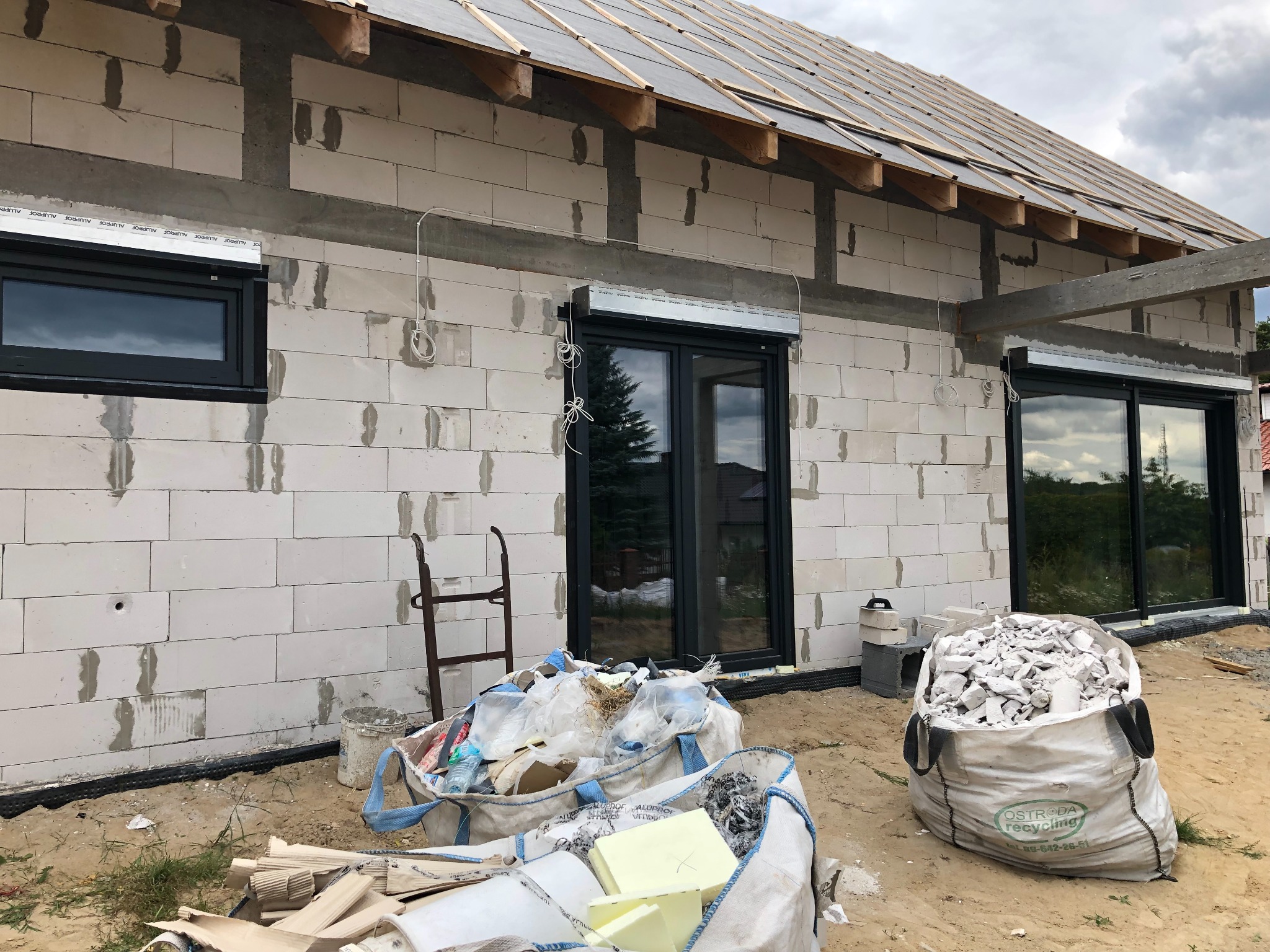 Nowy dom w trakcie budowy z zamontowanymi oknami i częściowo opuszczonymi osłonami okiennymi, widoczne worki z gruzem i materiałami budowlanymi, nieotynkowane ściany z białego betonu komórkowego.