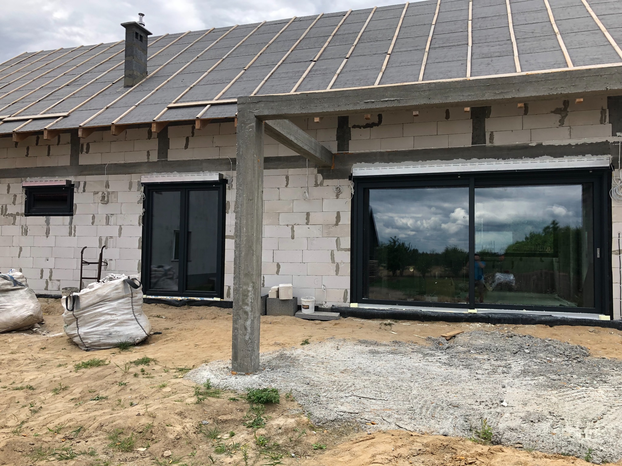 Surowy stan budynku jednorodzinnego z zamontowanymi oknami i częściowo opuszczonymi roletami zewnętrznymi, widoczne worki budowlane i rusztowanie, dach w trakcie budowy.