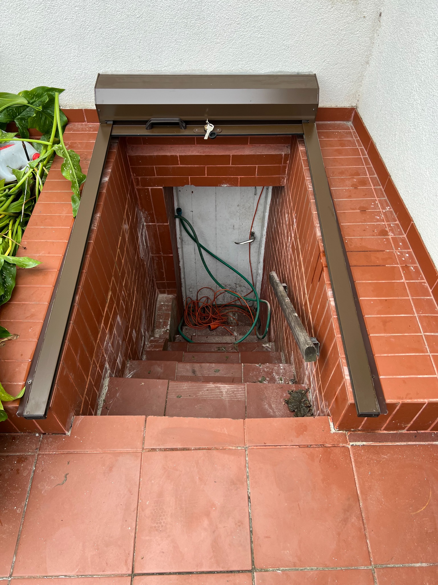 Zewnętrzne schody do piwnicy z brązową roletą zabezpieczającą, widoczne węże ogrodowe i kabel elektryczny wewnątrz.