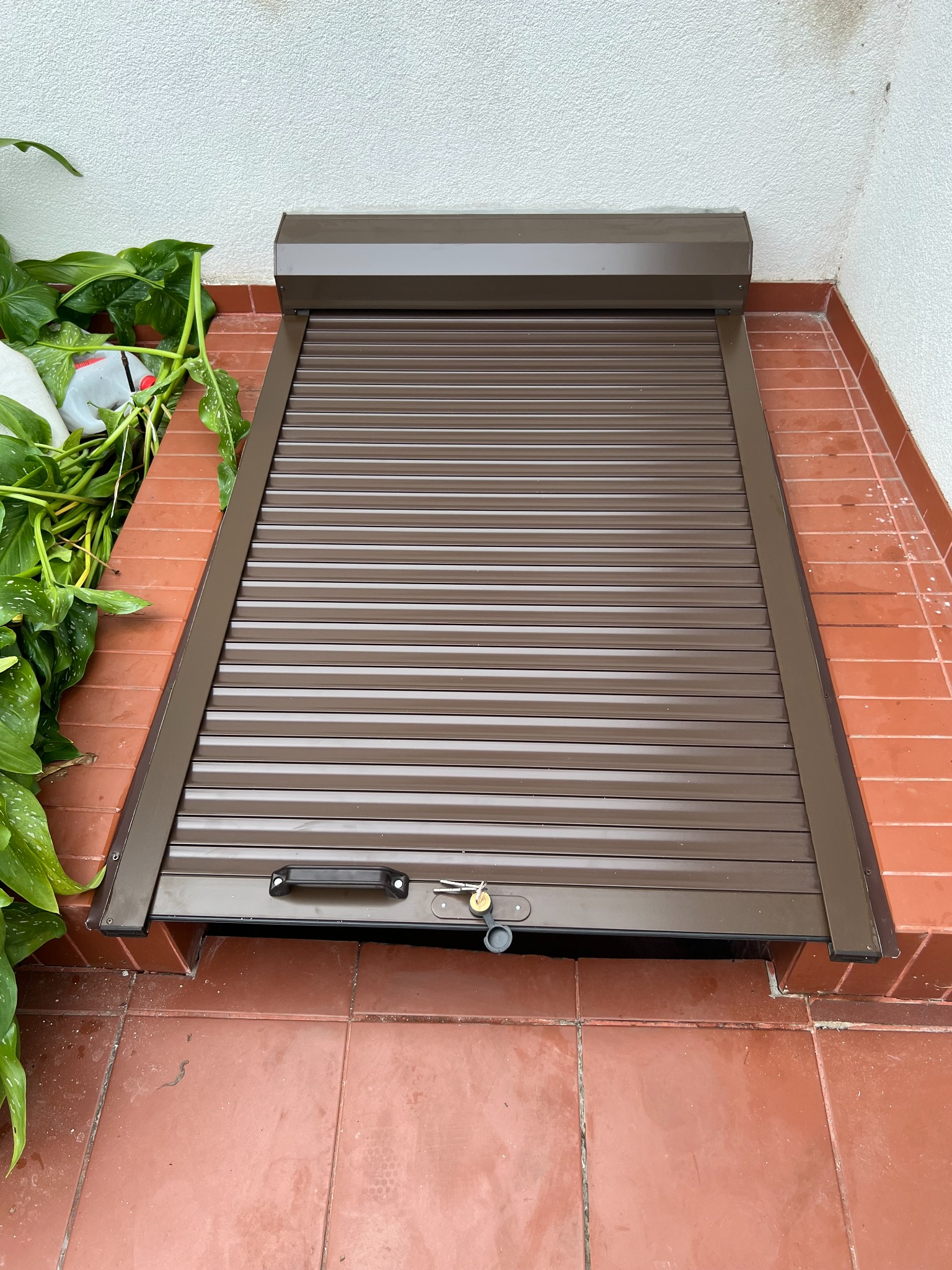 Zasłonięta brązowa roleta na poziomie podłogi, z kluczem w zamku, otoczona czerwoną terakotą i zielonymi roślinami.