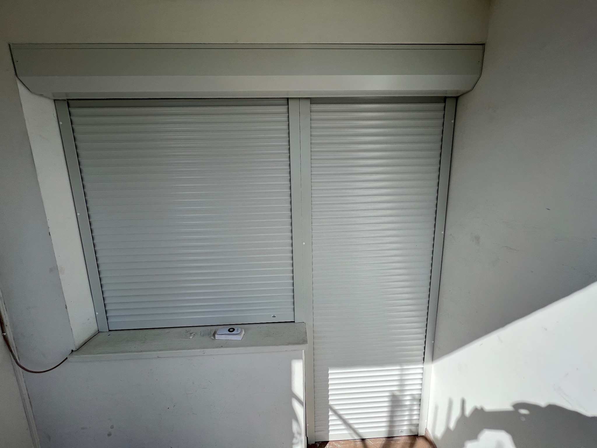 Zamknięte białe rolety zewnętrzne z widocznym sterownikiem na parapecie, wnęka okienna z plamami na ścianach