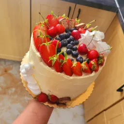 Okrągły tort z białym kremem, udekorowany świeżymi truskawkami, wiśniami, borówkami, bezami i kawałkami czekoladowych batoników, trzymany w dłoni na tle kuchennych szafek.