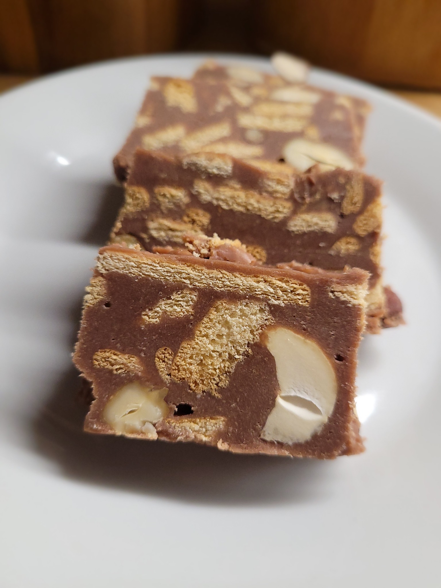 Trzy kawałki domowego ciasta czekoladowego z herbatnikami i orzechami na białym talerzu, widoczne nierówności i ręczne wykonanie.