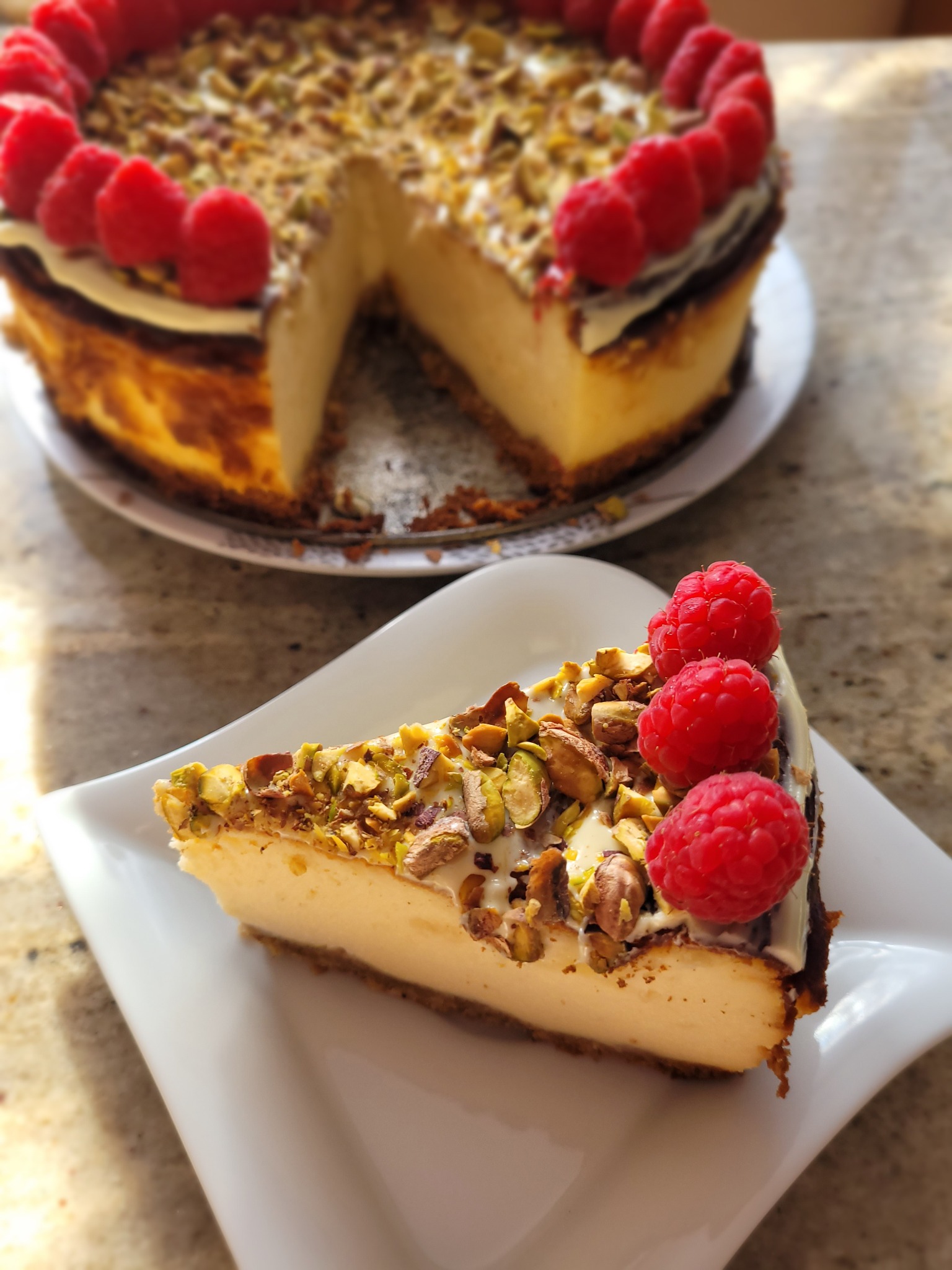 Kawałek sernika nowojorskiego z pistacjami i malinami na białym talerzu, w tle cały sernik z odciętym kawałkiem.