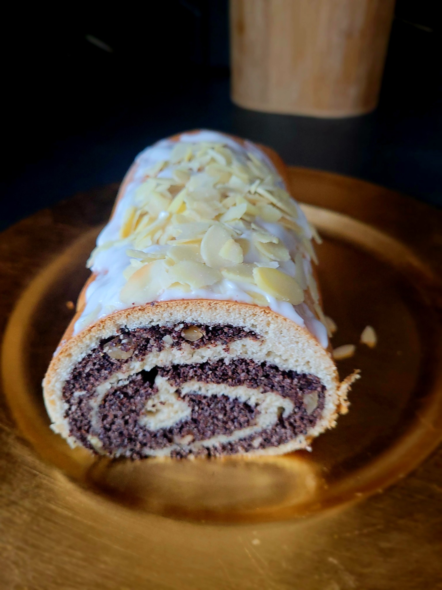 Przekrój makowca z lukrem i płatkami migdałów na złotej paterze, widoczne drobinki maku i orzechów w nadzieniu.