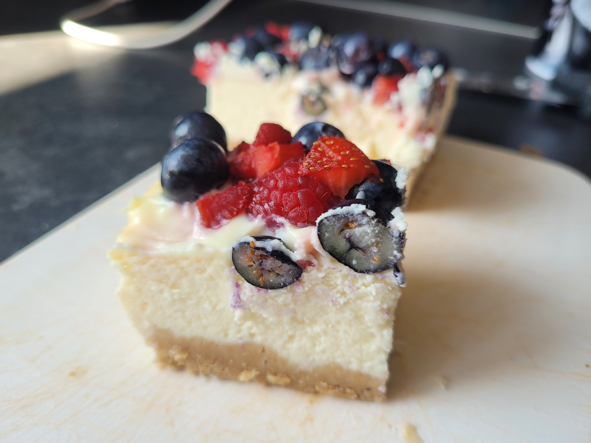 Kawałek sernika na kruchym spodzie, udekorowany świeżymi borówkami i truskawkami, leżący na drewnianej desce. Drugi kawałek widoczny w tle.