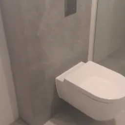 Nowoczesna toaleta podwieszana z szarą obudową stelaża podtynkowego i chromowanym przyciskiem spłukującym, na podłodze z jasnych płytek.