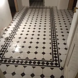 Podłoga wyłożona biało-czarną, błyszczącą ceramiką ułożoną we wzór karo z obramowaniem, widoczne niedokończone ściany i futryny drzwi.