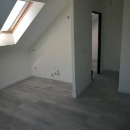 Poddasze po remoncie: szare panele podłogowe ułożone w jodełkę, białe ściany z widocznymi otworami instalacyjnymi i oknem dachowym, czarne obramowanie drzwi w głębi.