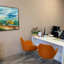 Wnętrze biura: obraz z motywem morza i łodzi, biurko z mapą świata, dwie pomarańczowe krzesła, roślina doniczkowa i materiały reklamowe.