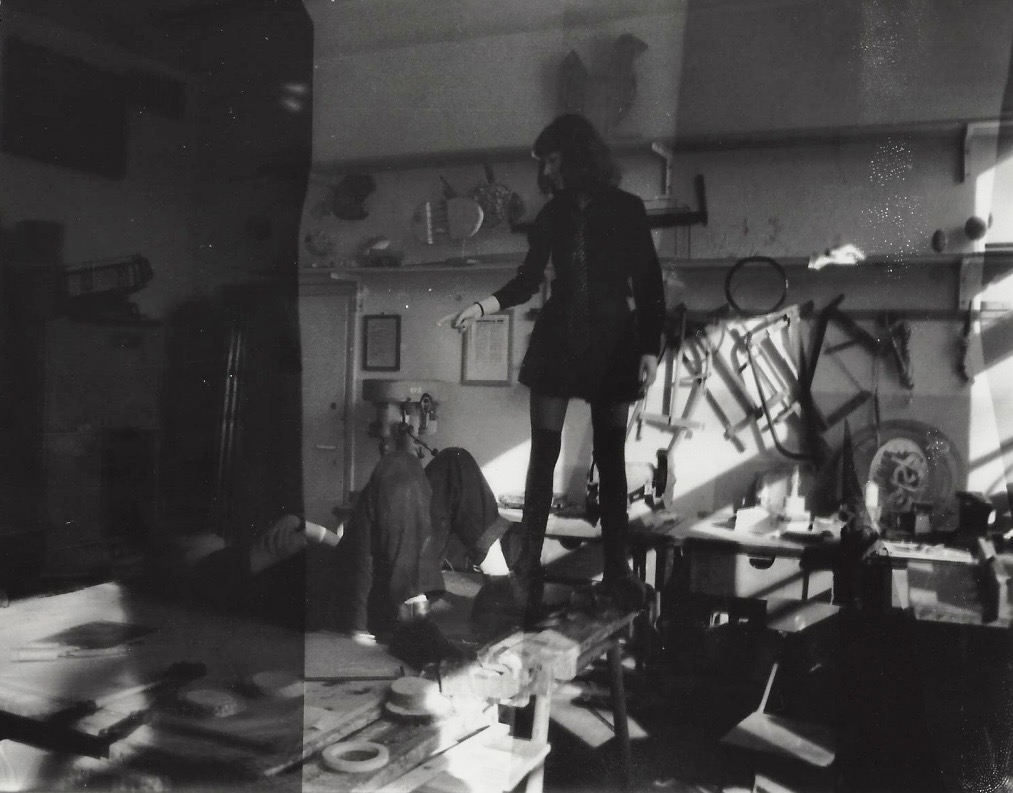 Czarno-biała fotografia analogowa przedstawiająca kobietę w ciemnej sukience stojącą na warsztatowym stole, wskazującą na ścianę z narzędziami w pracowni artystycznej.