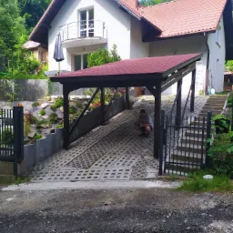 Nowoczesny dom jednorodzinny z białymi ścianami i czerwoną dachówką, z zadaszonym miejscem parkingowym wyłożonym ażurowymi płytami, w tle zieleń lasu, mężczyzna w kucki przy wjeździe.