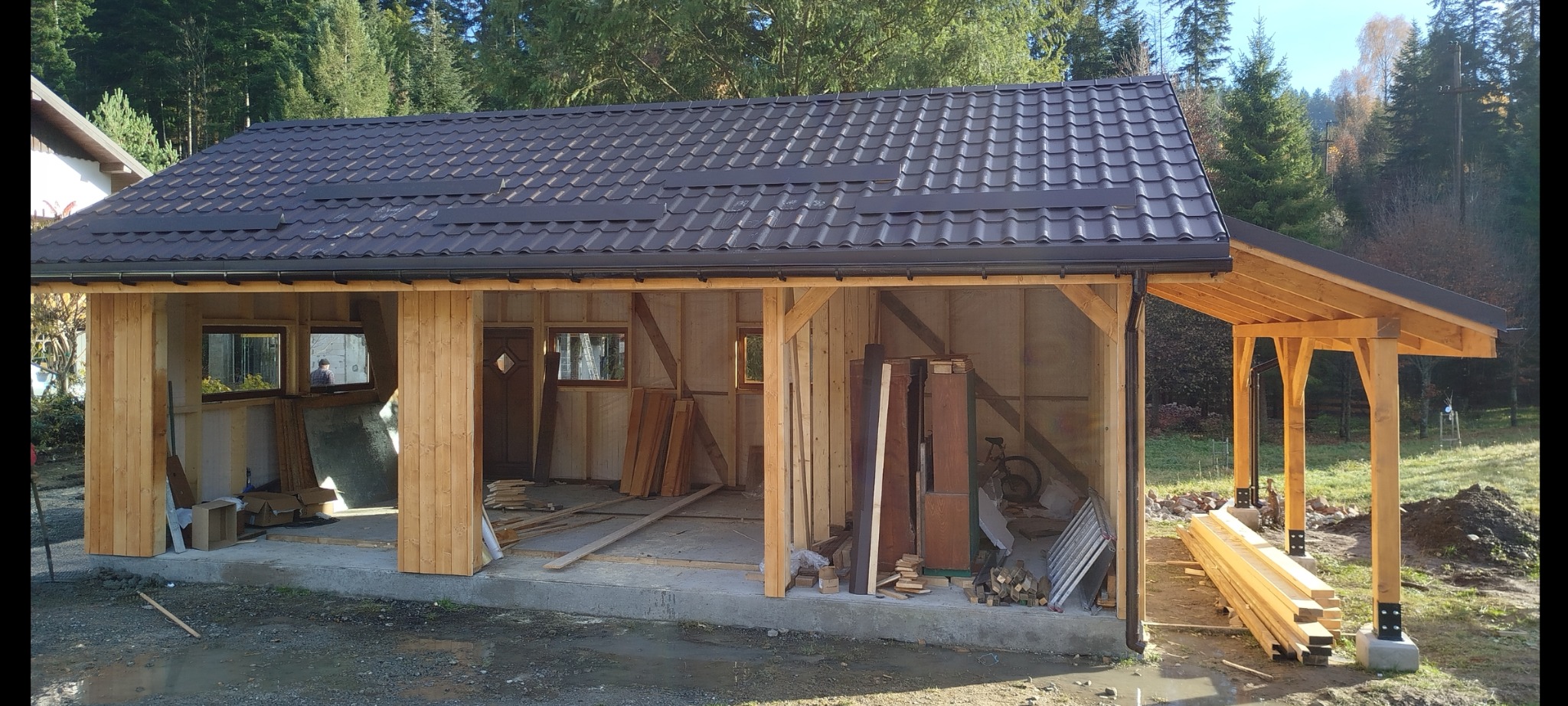 Drewniana konstrukcja altany w trakcie budowy, widoczne elementy szkieletu, dach z blachodachówki, drewniane słupy i przygotowane deski. Praca w toku.