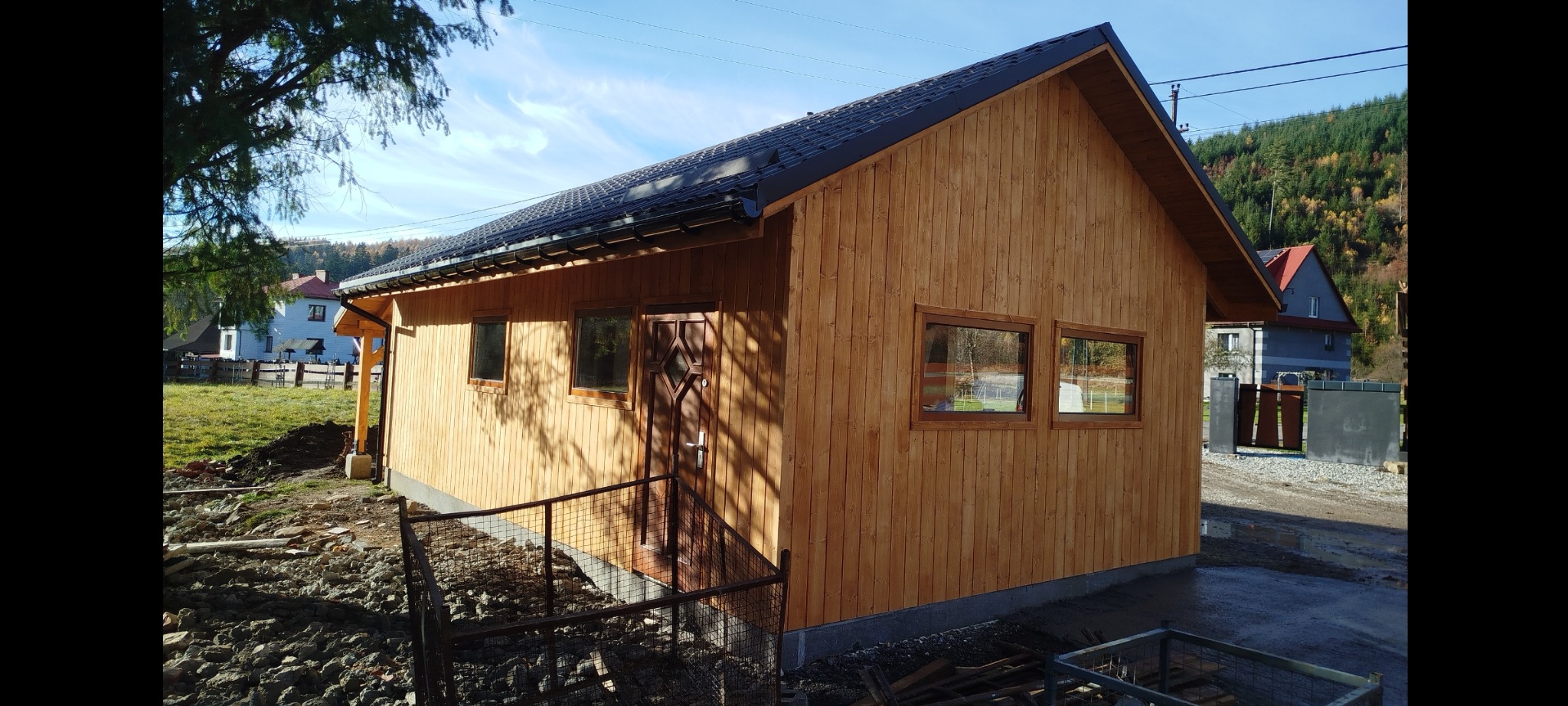 Drewniana altana z ciemnym dachem i oknami, otoczona ziemią i zielenią. Widoczne detale wykończenia i naturalne oświetlenie.