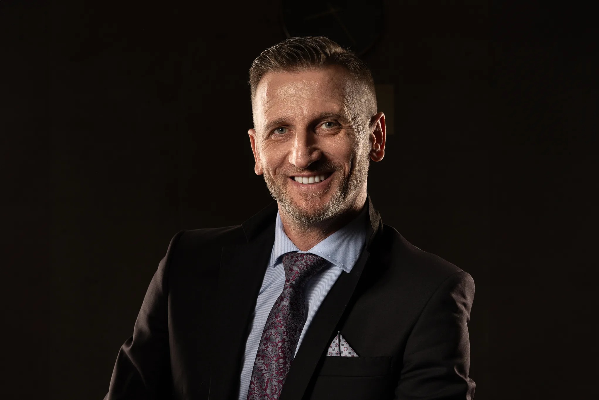 Portret uśmiechniętego mężczyzny w garniturze z wzorzystym krawatem i poszetką, wykonany w studio w Warszawie na ciemnym tle.