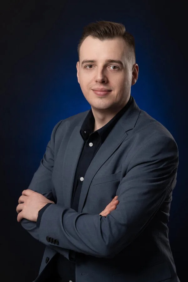 Portret mężczyzny w szarym garniturze i czarnej koszuli, pozującego na tle ciemnego, niebieskiego tła z rękami skrzyżowanymi na piersiach.