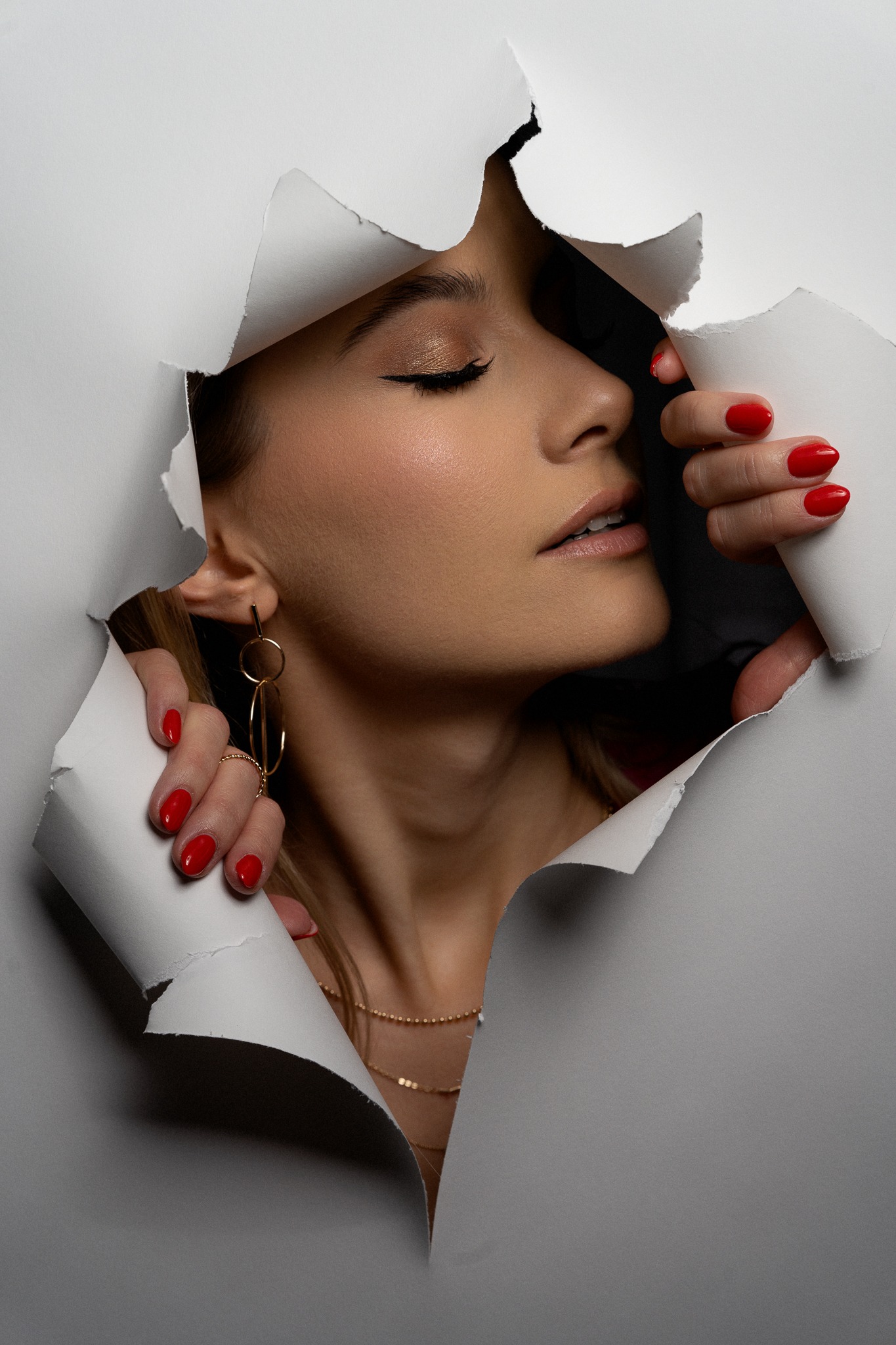 Portret kobiety z zamkniętymi oczami, wyłaniającej się z podartej kartki papieru, z widocznym złotym makijażem oczu, czerwonym manicure i złotą biżuterią