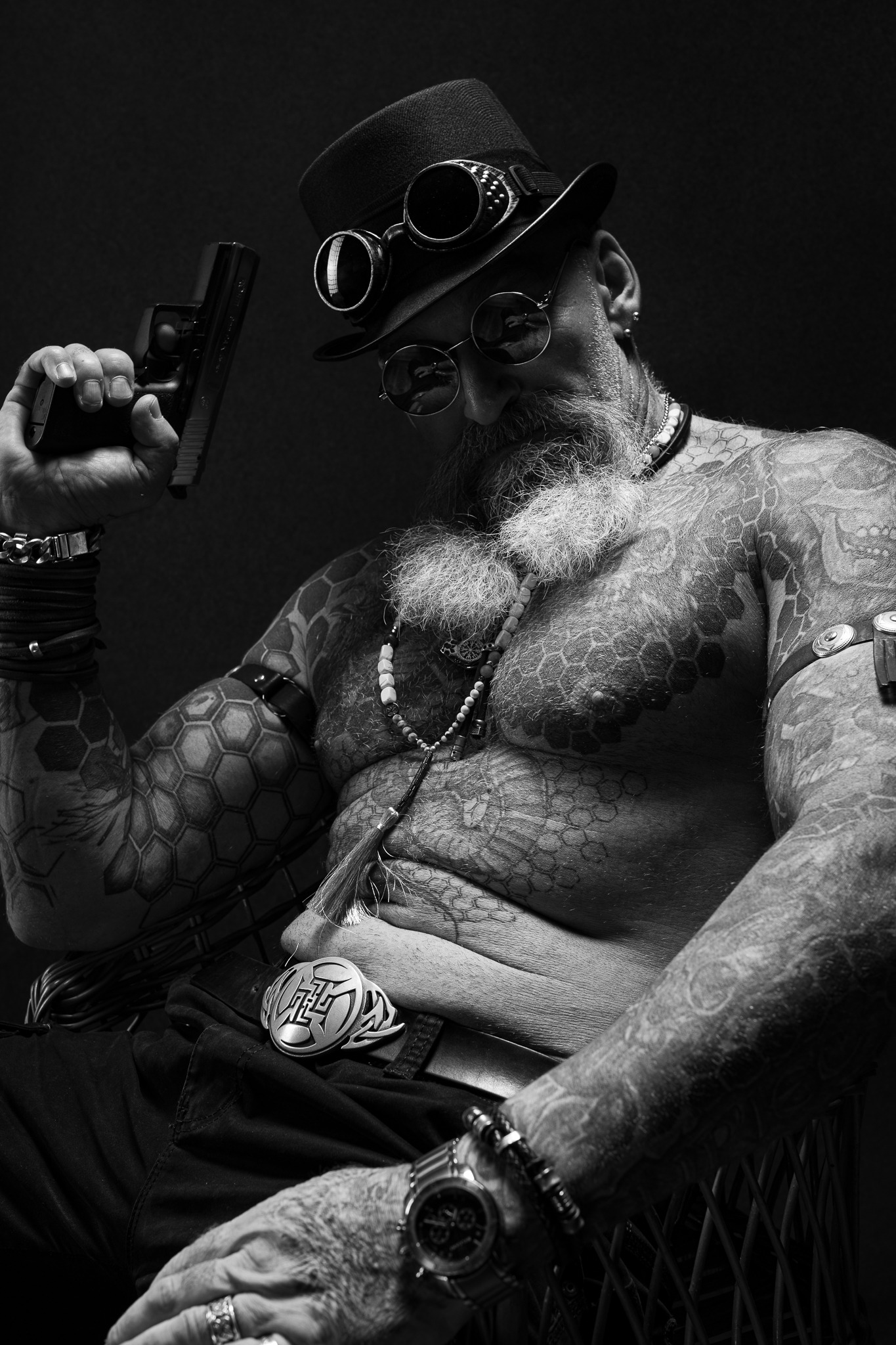 Czarno-białe zdjęcie mężczyzny z brodą i tatuażami, w kapeluszu, okularach steampunkowych i z pistoletem w dłoni, siedzącego na krześle.
