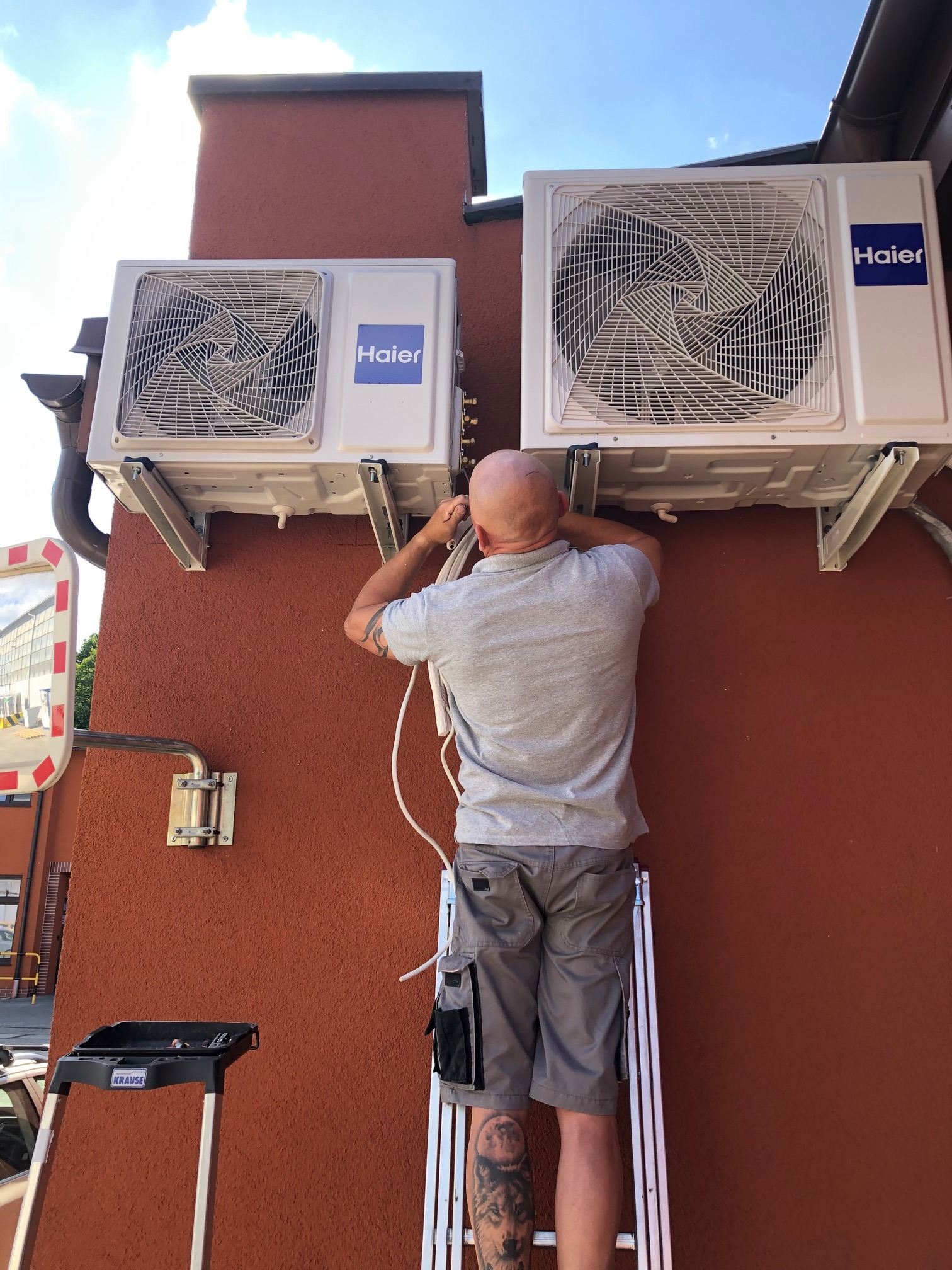 Instalator na drabinie montuje dwie jednostki zewnętrzne klimatyzacji Haier na brązowej ścianie budynku, widoczne przewody i narzędzia.