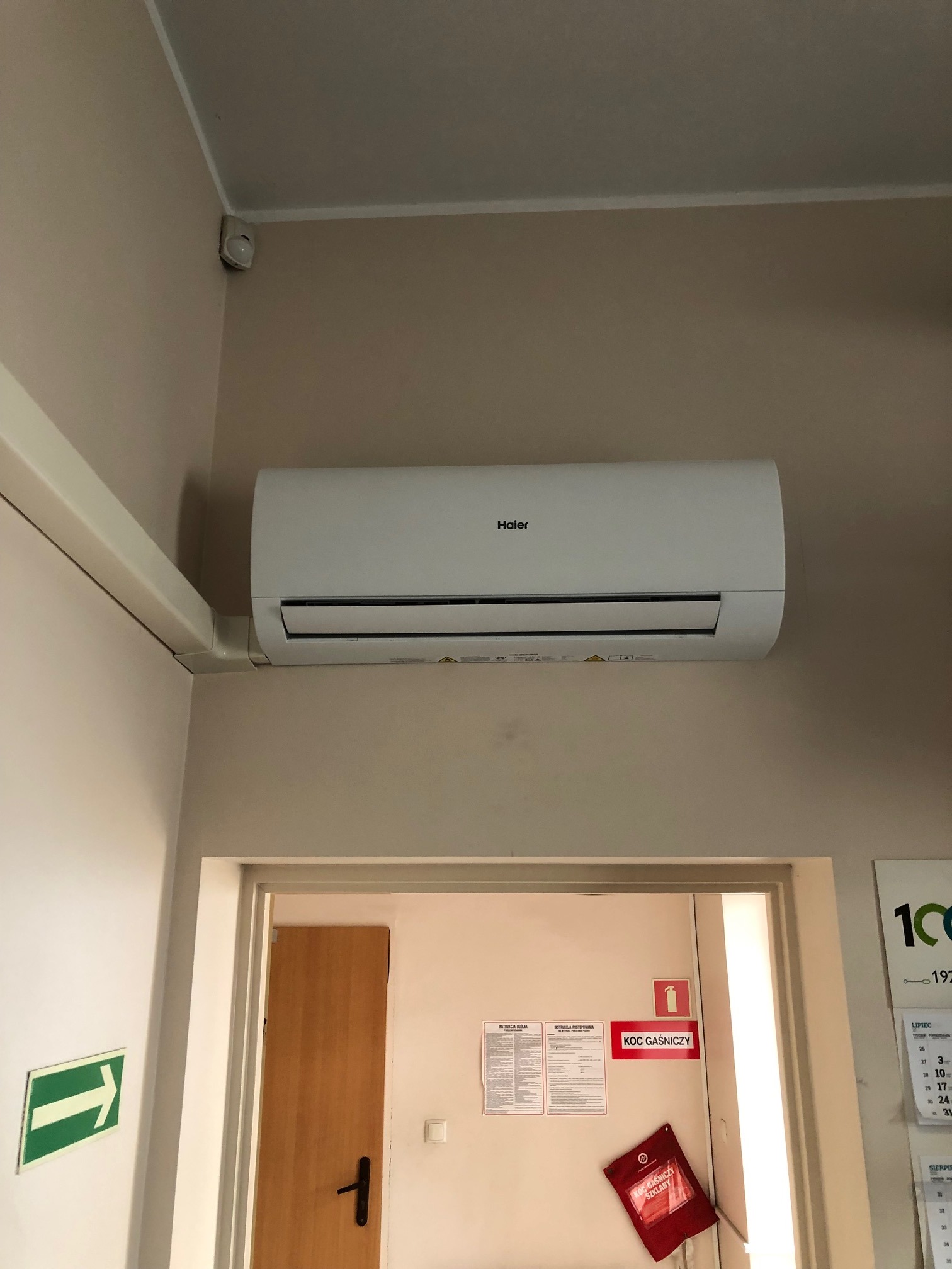 Biała klimatyzacja Haier zamontowana na ścianie w biurze, widoczny czujnik ruchu pod sufitem, instrukcja i koc gaśniczy na ścianie obok drzwi.