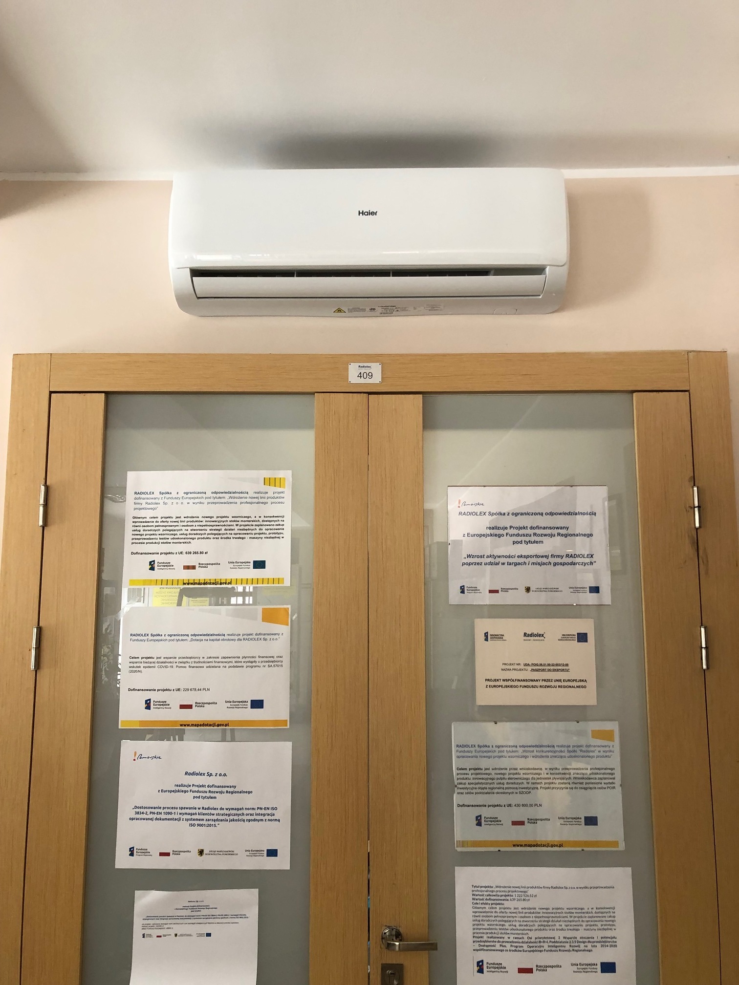 Biały klimatyzator Haier zamontowany na ścianie nad drewnianymi drzwiami z szybą, na których wiszą ogłoszenia firmy Radolex.