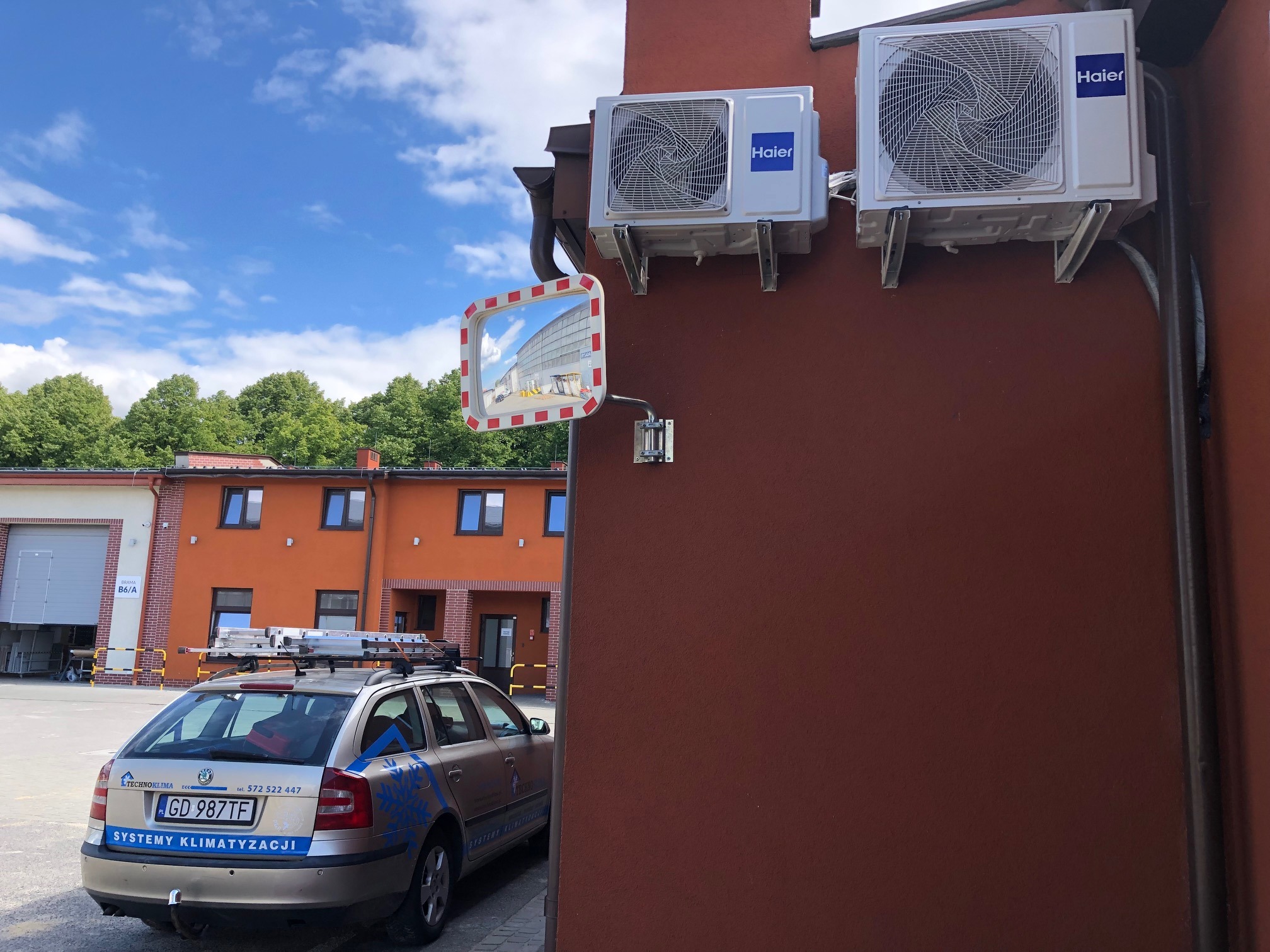 Dwa klimatyzatory Haier zamontowane na brązowej ścianie budynku. Poniżej widoczne lustro drogowe i samochód serwisowy z logo firmy i napisem 'Systemy Klimatyzacji' oraz drabiną na dachu.