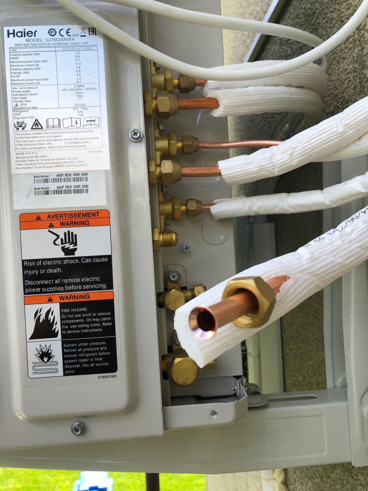 Zewnętrzna jednostka klimatyzacji Haier z widocznymi miedzianymi rurami w izolacji, etykietą z danymi technicznymi i ostrzeżeniami o ryzyku porażenia prądem i pożaru.