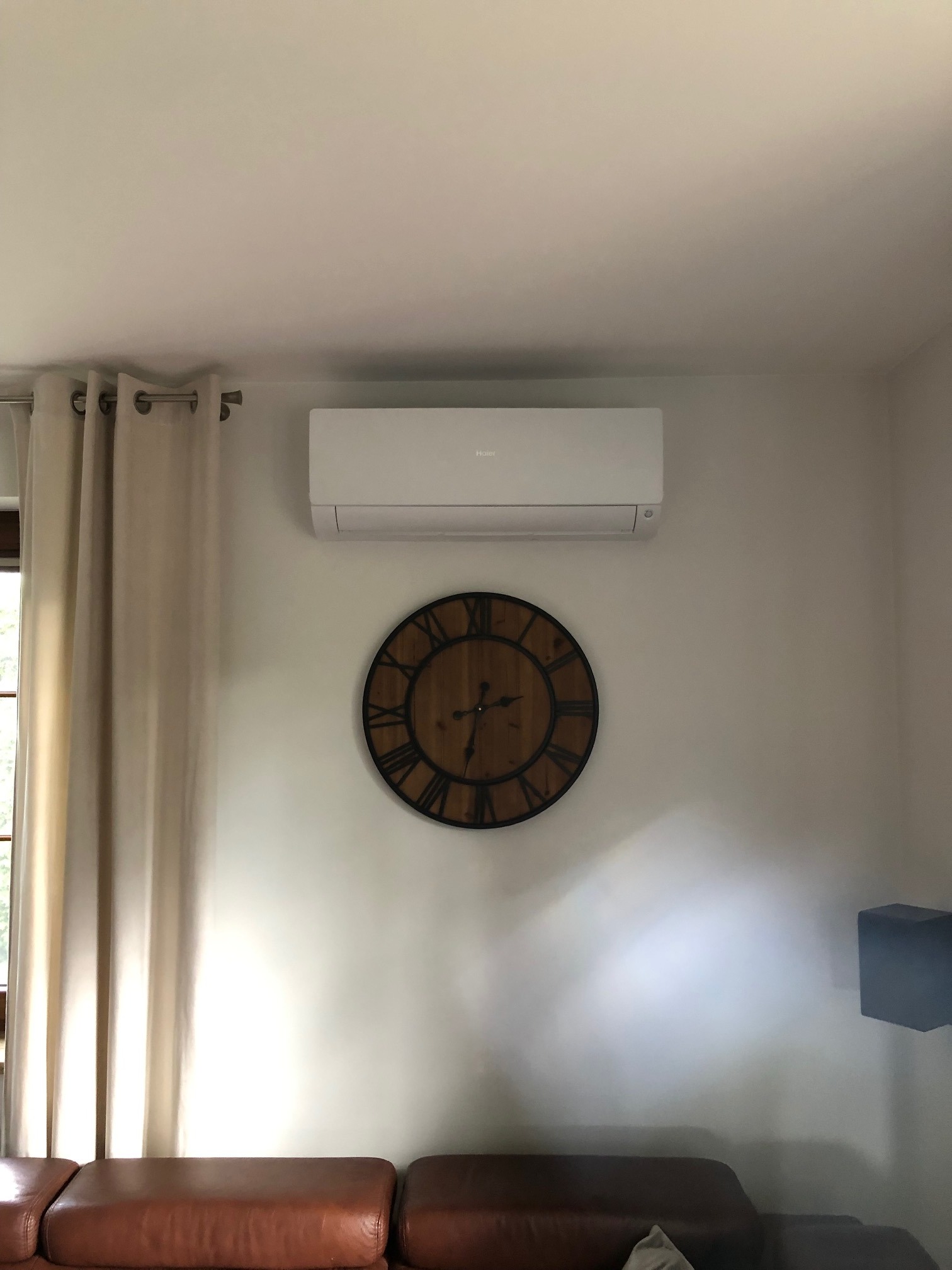 Biały klimatyzator marki Haier zamontowany na ścianie w salonie nad dużym zegarem z rzymskimi cyframi i brązową skórzaną sofą.