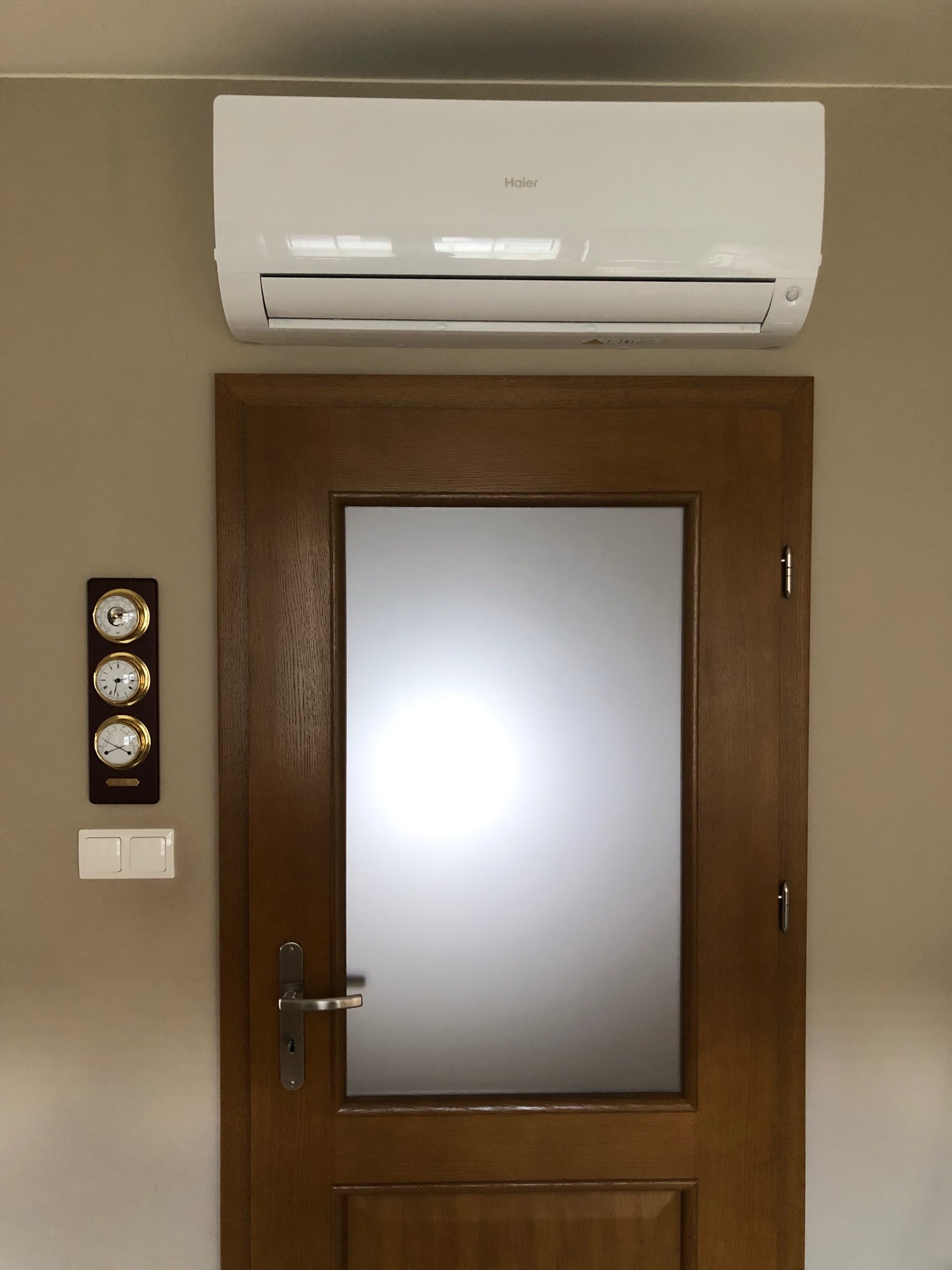Biały klimatyzator Haier zamontowany nad drewnianymi drzwiami z matową szybą, obok panel z trzema zegarami i włącznikiem światła na beżowej ścianie.