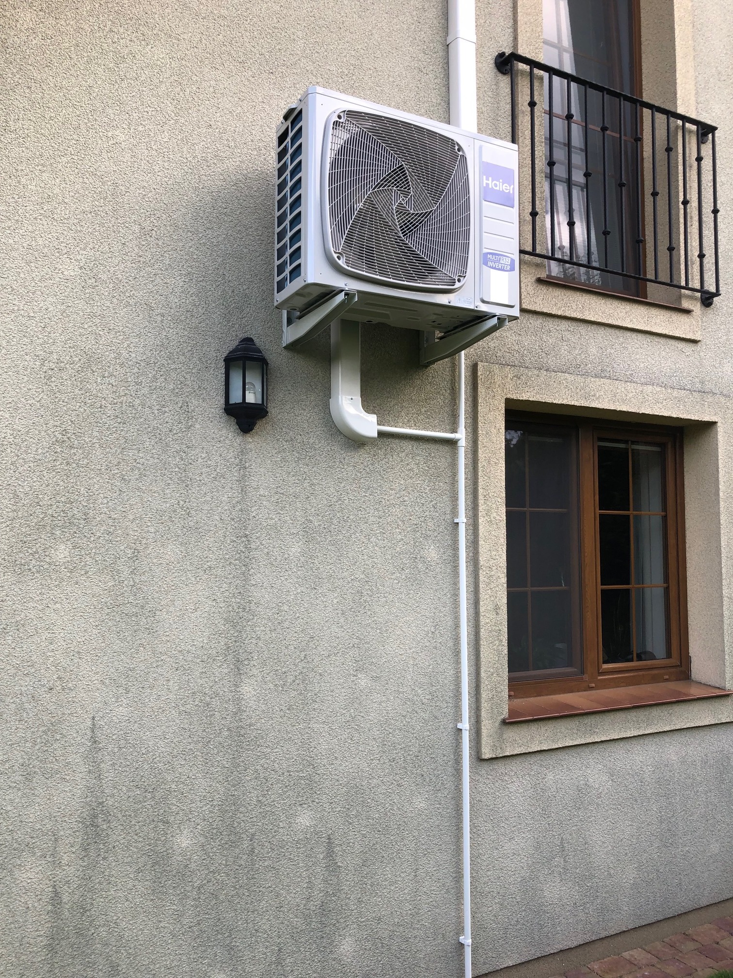 Zewnętrzna jednostka klimatyzacji Haier zamontowana na elewacji domu, widoczna lampa zewnętrzna i okno z ozdobną balustradą.