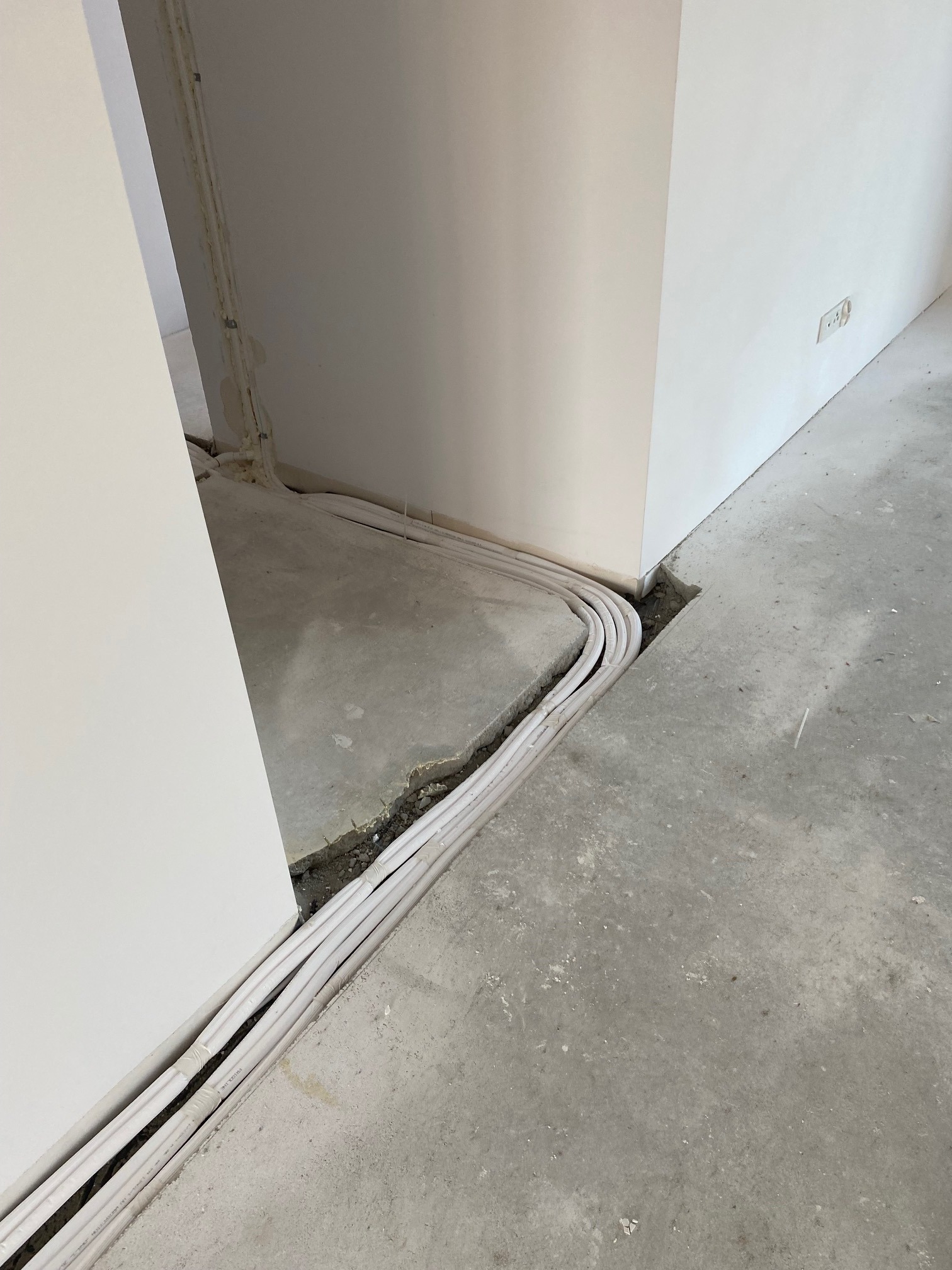Białe przewody ułożone w wyżłobieniu w surowym betonowym podłożu, wzdłuż ściany w trakcie remontu. Widoczne gniazdko elektryczne na ścianie.