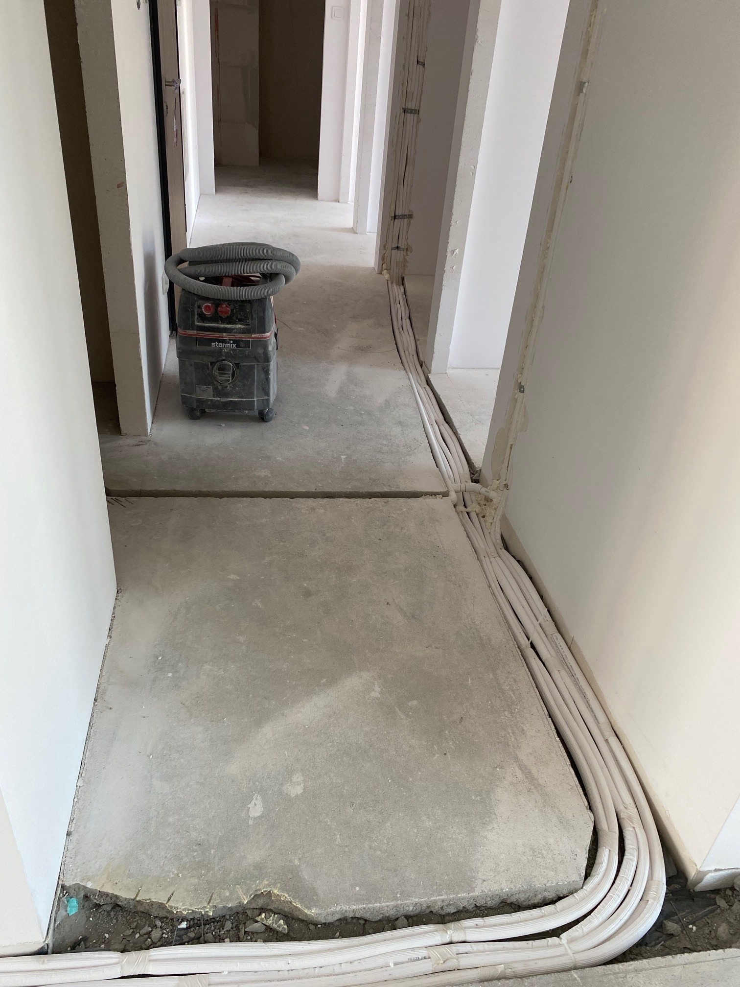 Białe rury instalacyjne ułożone w bruzdach w podłodze i na ścianie, widoczny odkurzacz przemysłowy Starmix w surowym wnętrzu budynku.